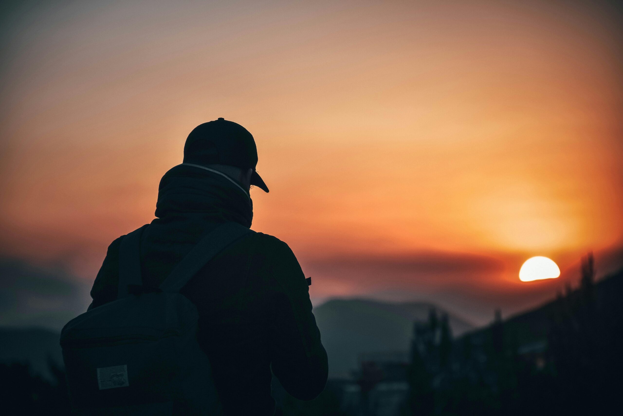 A man with his back to us wearing a peaked cap looking over a sunset landscape.